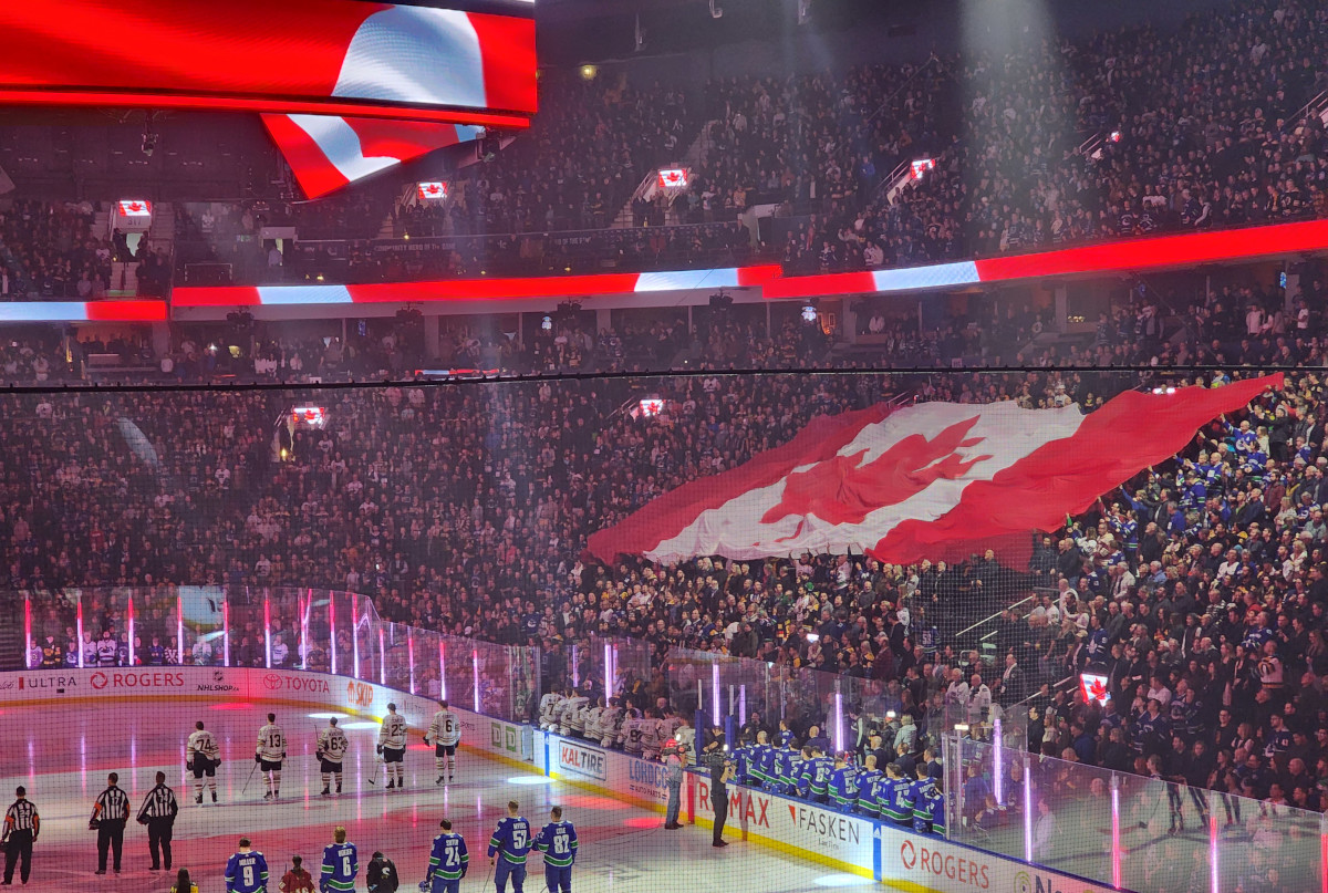 An enormous Canadian flag held up by a full stadium audience watching hockey players and referees lined up on the ice.