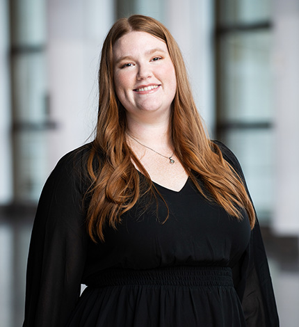Light-skinned smiling adult with long wavy auburn hair, a necklace, and a black top.