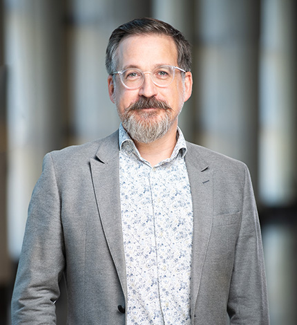 Light-skinned adult with short brown hair, a brown moustache, a greying beard, a grey blazer, and a patterned white dress shirt.