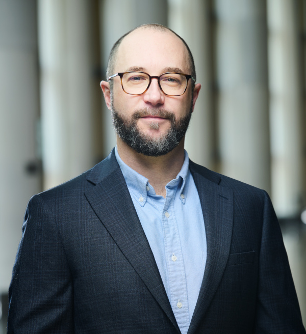 Light-skinned adult with a balding head, a brown moustache and beard, round glasses, a dark blue jacket, and a light blue shirt.