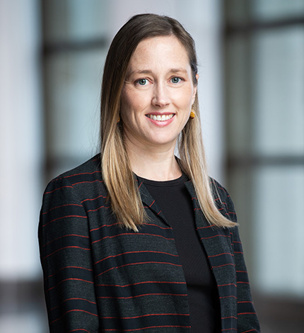 Light-skinned smiling adult with long straight brown hair, earrings, a striped blazer, and a black top.