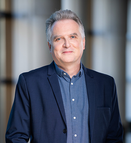 Light-skinned adult with short, greying hair, a dark blue blazer, and a blue button-down shirt.