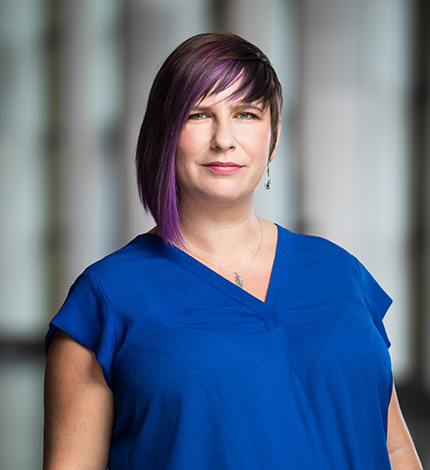 Light-skinned adult with asymmetrical purple hair, silver earrings and necklace, and a blue top.